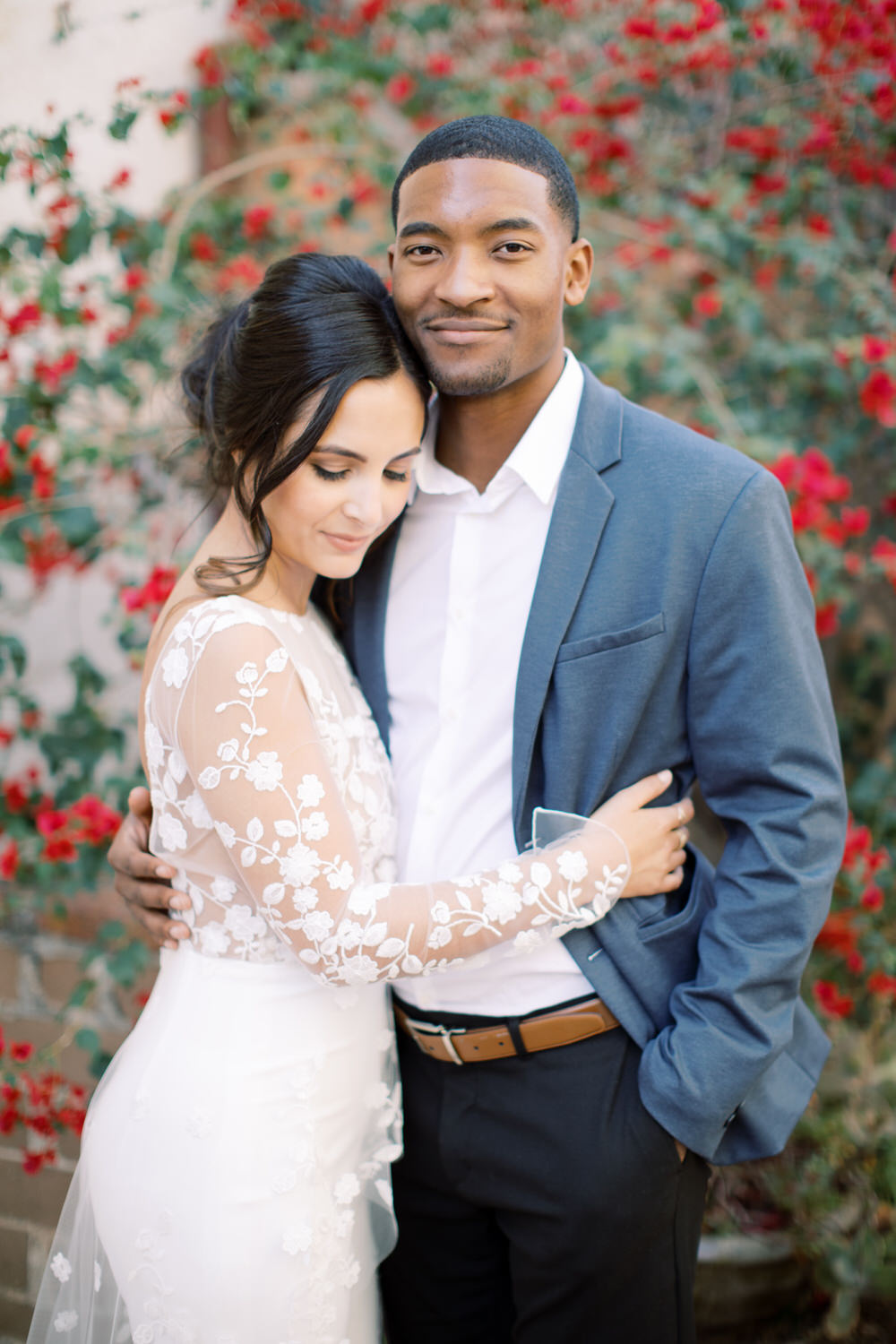 black man in blue suit embracing his fiancee in wedding dress against red flowers wall